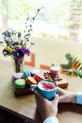 Girl hand hold a blue cup of morning cappuccino coffee. French macarons and roses petals on wood desk.