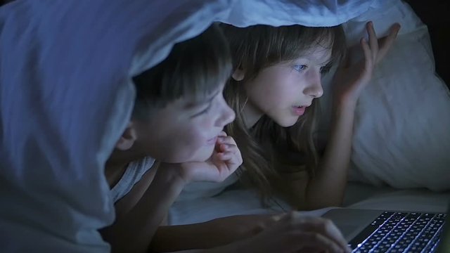 A boy and a girl are playing on the computer and lying on the bed under the blanket at night.