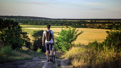 A tourist man stands holding a bicycle in front of an opening landscape under a blue sky