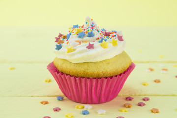 Single vanilla sponge birthday cupcake frosted and coated with star candy sprinkles on pale yellow background