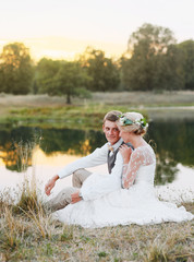 Fototapeta na wymiar Just married loving hipster couple in wedding dress and suit on green field in a forest at sunset. happy bride and groom sitting in the summer meadow. Romantic Married young family.