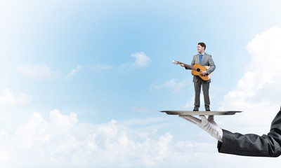 Businessman on metal tray playing acoustic guitar against blue sky background