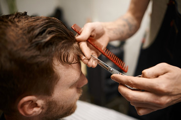 Time for a new haircut. Handsome stylish young Caucasian bearded man came to barbershop for hairdo. Unrecognizable hairstylist holding scissors and comb while cutting or trimming client's hair
