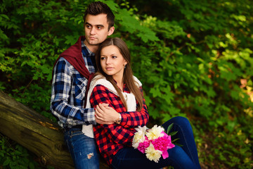 Stylish young couple in shirts and jeans while walking in the forest. A beautiful girl with her handsome boyfriend in a hug sitting on a fallen tree.