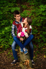 Stylish young couple in shirts and jeans while walking in the forest. A beautiful girl with her handsome boyfriend sitting on a fallen tree.