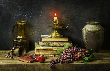 Classic still life with sweet red grapes placed with vintage books, illuminated candle,old jar,pipe...