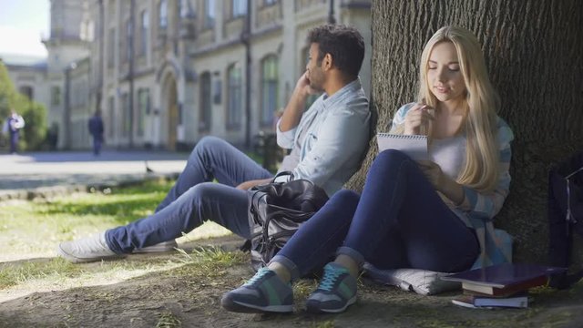 Multiracial guy talking on cellphone under tree, girl taking notes in notebook