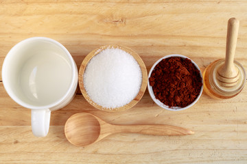 wooden honey dipper in honey bottle and sugar in wooden bowl and cocoa in ceramic bowl and blank ceramic cup on wooden table, top view