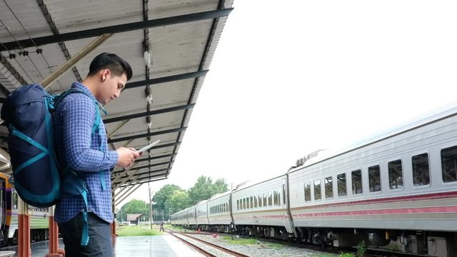 Young Asian Man With Backpack Standing On Platform At Train Station. Backpacker Or Traveler Using Digital Tablet While Waiting For Train. Journey, Trip, Travel Concept