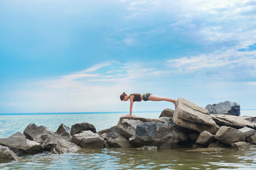 Girl practicing yoga on the rocks against the blue sky and the azure sea. Woman stands on a stone in a plank posture.
