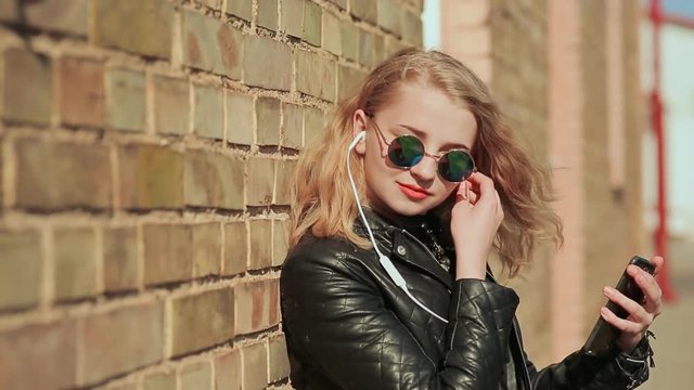 Stylish young blonde in sunglasses and leather jacket listening to music on bluetooth headphones in a mobile phone. Enjoy music. Spring. Background of a brick wall.