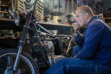 Fototapeta na wymiar Man smoke during a break of repairing old motorcycle in workshop