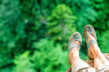 Woman feet in sandals hung from mountain rock above precipice on green forest trees background