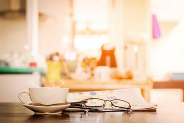Closeup cup of hot cappuccino coffee with newspaper and spectacles on the wood table with blur coffee shop background-warm tone.