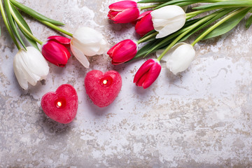 Two red burning candles in form of heart and red  and white tulips flowers on grey background.