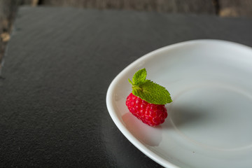 Ripe juicy raspberries on a plate on dark background. Fresh green mint leaves.