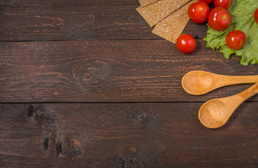 Two wooden spoons on a dark wood background. Vegetables, red cherry tomatoes, salad leaves and cripsbreads vitamins delicious at the right side. Vegeterian or vegan background.
