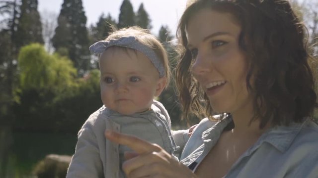 Happy Mother Holds Cute Baby Girl And Points To Something In Distance In Park On Sunny Day