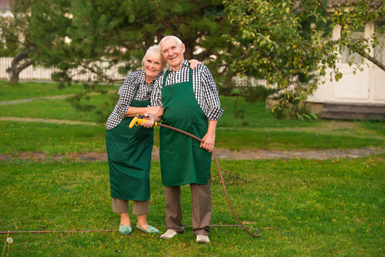 Senior Gardeners Couple, Water Hose. Smiling Man And Woman Outdoors. Garden Improvement Ideas.