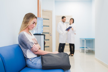 Young woman filling nervous sitting at hospital corridor waiting while two doctors talking on a backgroud