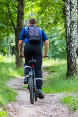 Fototapeta na wymiar A seventeen-year-old boy, a teenager, rides a bicycle through the forest in the mountains. He is wearing a blue T-shirt and black sports pants, with a backpack behind his shoulders.
