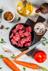 Meat, beef. Fresh raw chopped goulash, beef cubes in a bowl. Spices (salt, pepper), tomatoes, garlic, onions. On a white marble table, with a fork for meat and a knife. Copy space top view