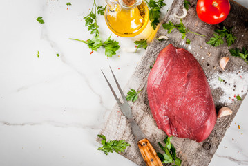 Cooking beef fillet tenderloin. Large whole piece of veal fillet on an old cutting board with a fork for meat, spices (salt, pepper, parsley, garlic, onions, tomatoes). Copy space top view