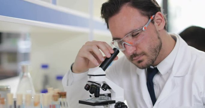 Male Scientist Working With Microscope Looking At Test Tube In Laboratory Wearing White Coat And Protective Glasses Slow Motion 60