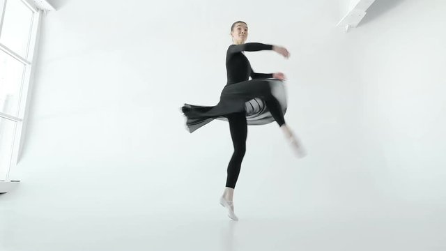 Attractive brunette ballerina in white pointe and black dress dancing in a white dance-hall near the big window 20s 1080p slow motion
