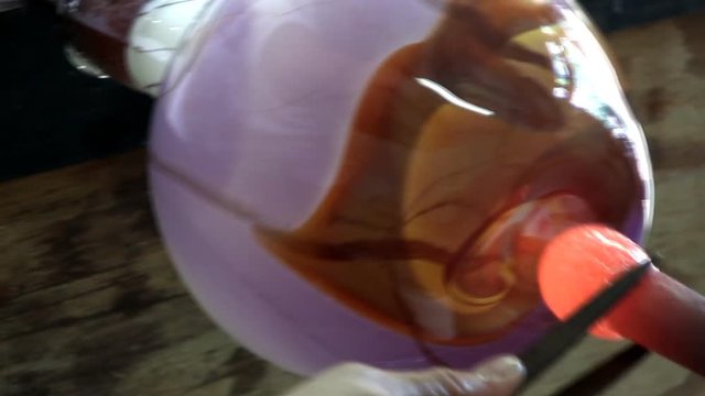 A punty being attached to base of multicolored vase.  Glass artist uses tweeters to help steady and center pipe.  An air hose is used to cool the punty after attachment