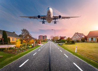 Naklejka premium Beautiful cityscape with passenger airplane is flying in the sunset sky above the asphalt road through the town with houses and courtyards at sunset in Netherlands. Landing airplane. White aircraft