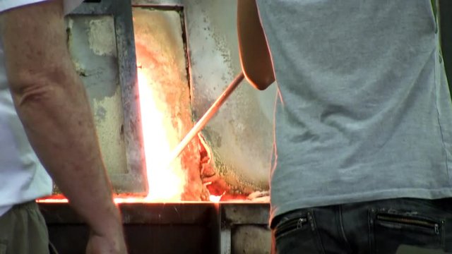 Young teenage girl apprentice grabs gather of glass from furnace as a master glass gaffer advises her