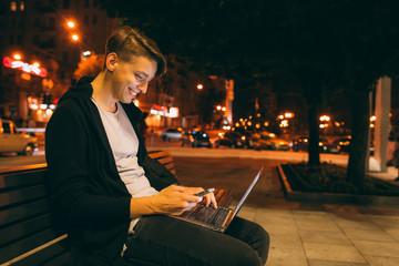 Young guy chatting in city street in night time, blurred lights background, free space. Smiling guy reading message on smartphone, while he working on laptop outside.