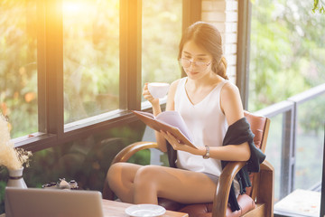 Young pretty Asian girl sitting near opened window drinking coffee and reading book enjoys of rest,women working university project with generic design laptop. Analysis marketting plans,vintage color