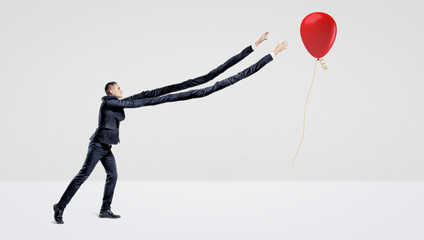 A businessman with extremely long arms trying to catch a red balloon with a gift gold ribbon.