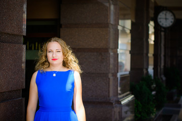 Blond hair curly woman plus size European appearance in blue retro dress walk in city at sunny warm day with good mood