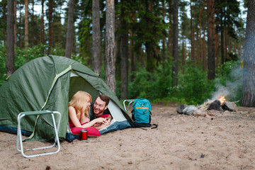 Beautiful couple lying in a tent, camping on the background of fire. Next to folding chairs, camping a mug of warm soup and a Hiking backpack.