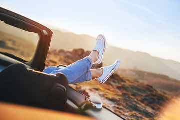 Pretty woman rests and pushes her sneakers out of the convertible to enjoy sunset - obrazy, fototapety, plakaty