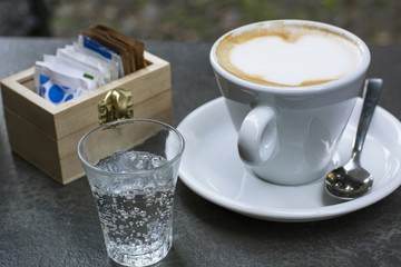 One white cup of cappuccino with small glass of water on the grey table