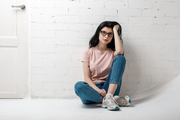 Portrait of smart beautiful brunette girl in eyeglasses with natural make-up sitting near white brick wall.