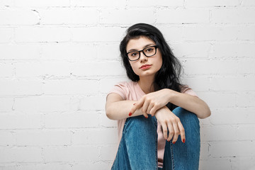 Portrait of smart beautiful brunette girl in eyeglasses with natural make-up sitting near white brick wall.