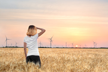 A girl in a wheat field looks at windmills, dawn