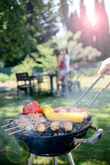 close up of a barbecue grill with meat and sausages cooking during summer garden party with people in background