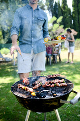 close up of a barbecue grill with meat and sausages cooking during summer garden party with people in background