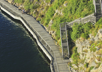 Beautiful summer season specific photograph. Pier/walkway/harbor/wooden path close to the ocean. Photo taken from above. Green vegetation, plants and rocks next to the wooden walkway. - 163011864