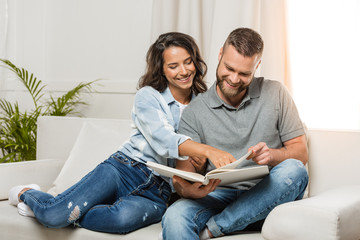 young smiling couple looking at photo album and sitting on sofa at home - obrazy, fototapety, plakaty