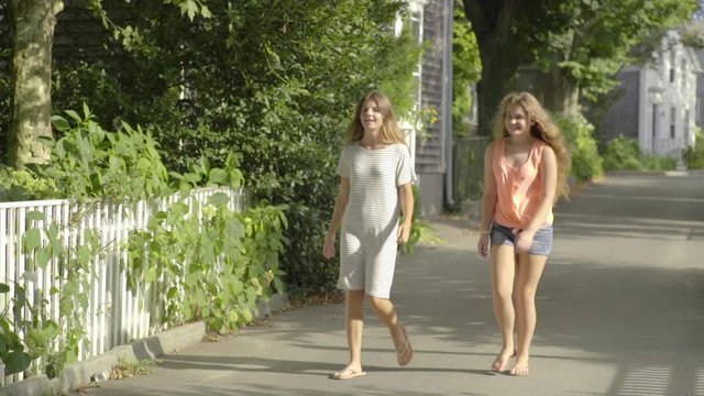 Two Teenage Girls Walk Down Middle Of Road In Beautiful Neighborhood