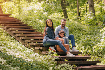 young smiling interracial family with picnic basket sitting on wooden stairs in sunny forest