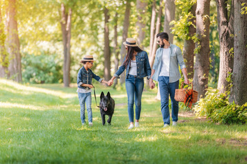 young happy interracial family with dog holding hands and walking in sunny forest