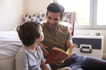 Father And Son Siting On Bedroom Floor Reading Book Together